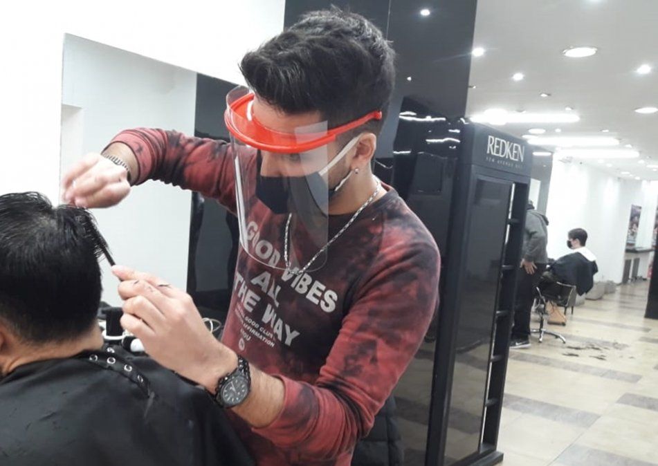 La nueva normalidad de las peluquer&iacute;as en la cuarentena. David, de la zona c&eacute;ntrica de Lomas, trabaja con barbijo y mascarilla protectora. Muy lejos en el fondo, otro cliente con barbijo.